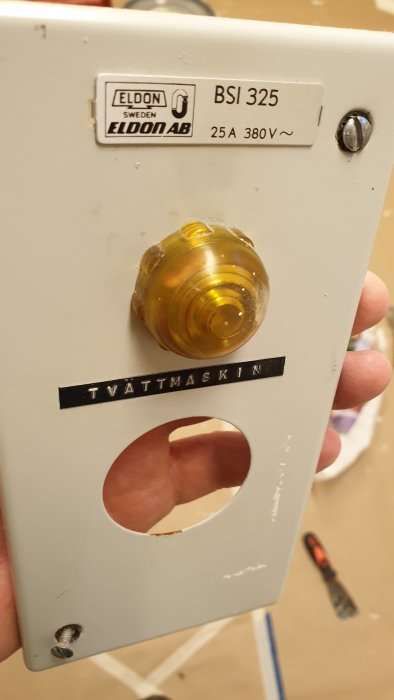 Eldon strömbrytare med gul plastlins, etikett "ELDON AB", text "TVÄTTMASKIN" och synlig hålighet där linsen saknas.