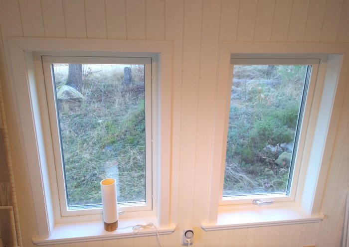 Två nyinstallerade fönster med vita smygar och foder i ett ljust rum, utsikt mot natur.