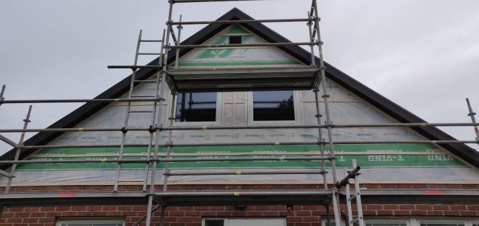 Husfasad under renovering med ställningar, visar isoleringsmaterial, fönster och vindskydd.