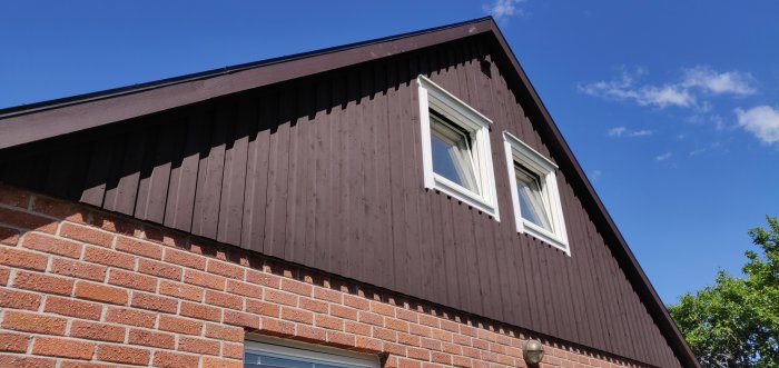 Hus med en kombination av tegel och mörka trätavlor, två vita fönster under snedtak, mot klarblå himmel.