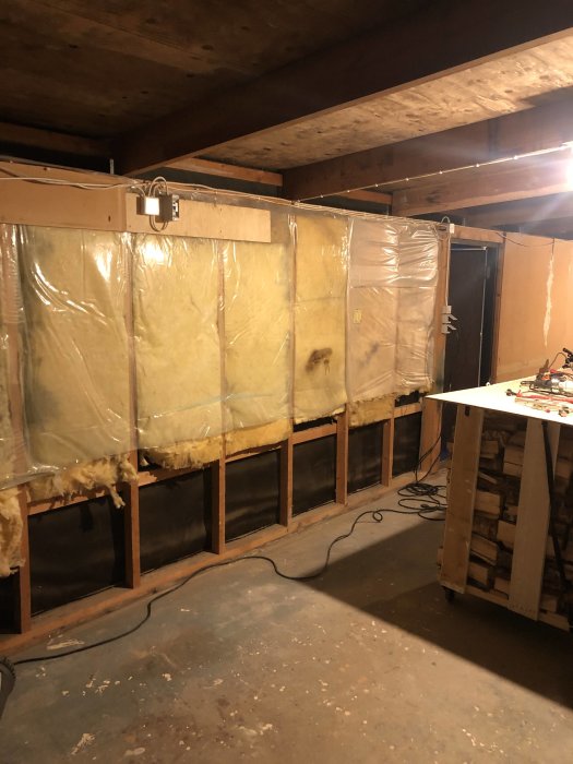 Ofullständigt isolerat garage med synliga träreglar och hängande isoleringsmaterial, samt en arbetsbänk.