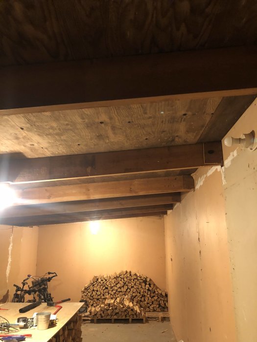 Interiör av ett kallgarage med synliga träbjälkar i taket och staplad ved mot väggen.