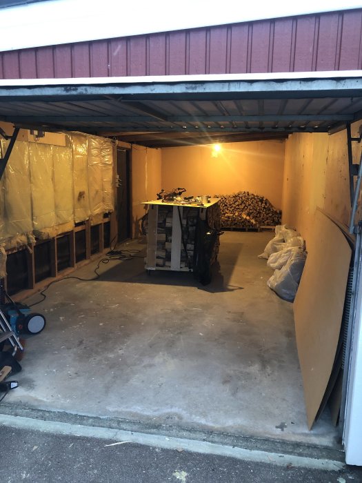 Öppet garage med synliga träreglar och isolering, plastad vägg och staplad ved i hörnet.