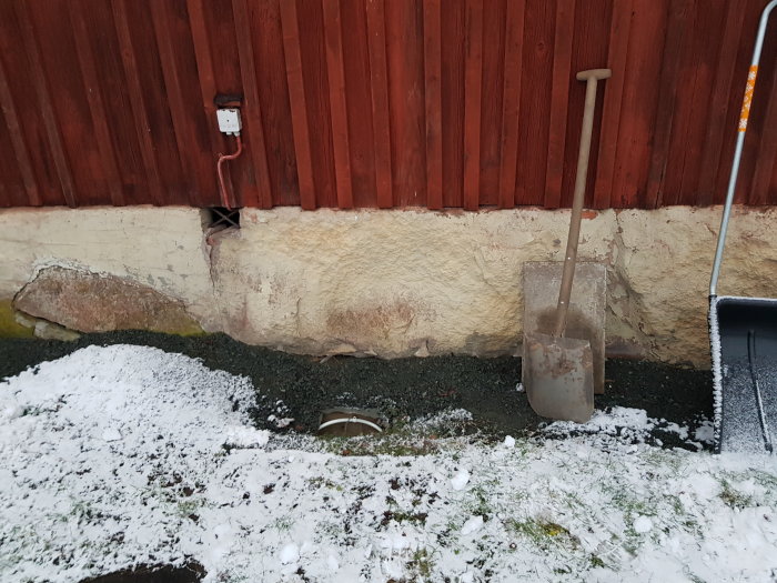 Spad och öppen fiberbrunn vid husvägg med nysprutad täckfärg och snötäckt mark.