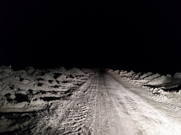 Snötäckt väg med spår och plogade snöhögar vid sidan om nattetid.