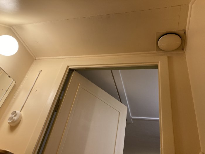 Vy över en toalettdörr med en tallriksventil högt upp på väggen, tänkt för frånluft.