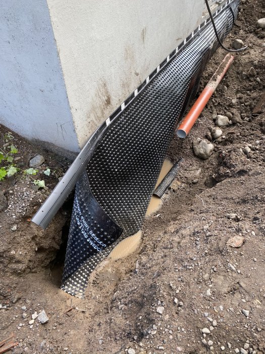 Installation av platonmatta längs fundament, med dräneringsrör och grus synligt i utgrävd gräns.