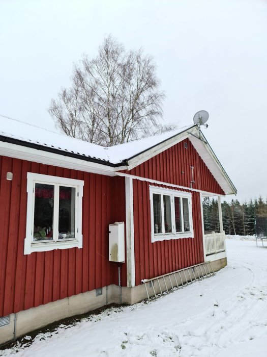 Rödmålat hus med vita fönster och snö på marken, fuktfläck på väggen bredvid elskåpet.