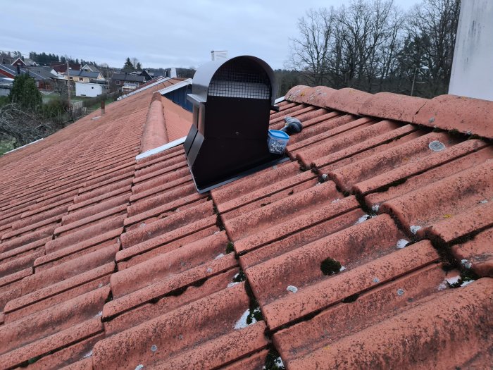 Nyinstallerad takfläkt på ett rött tegeltak med bostadshus och träd i bakgrunden.