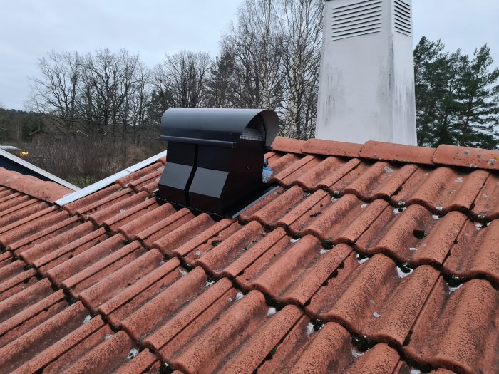 Nyinstallerad svart takhuva på röd tegelpannor med bakgrund av vit skorsten och träd.
