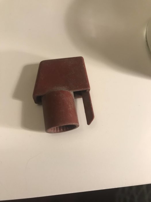 En brun plastdel, troligen en komponent till möbler, ligger på ett vitt underlag.