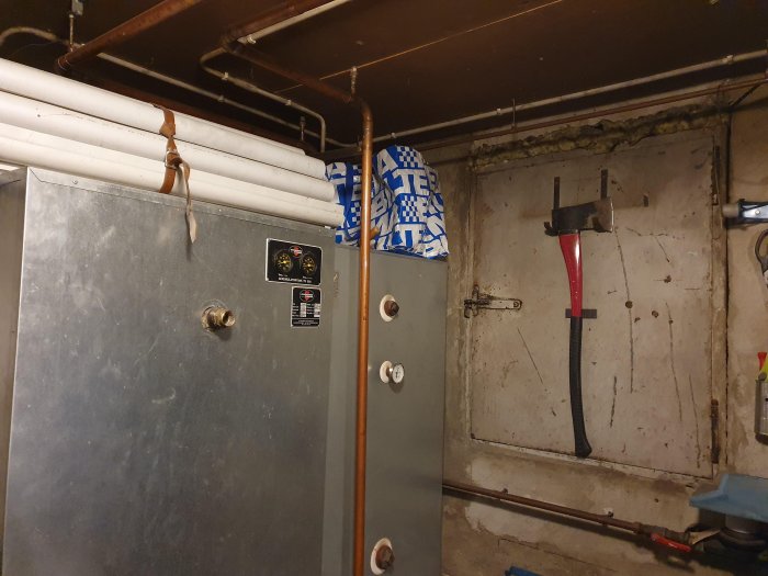En grå uppvärmningspanna i ett pannrum med kontrollmätare, kopplade rör och en upphängd yxa på betongväggen.