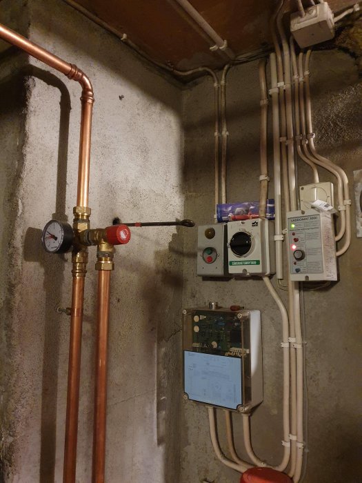 Värmesystem med kopparledningar, manometer, röda ventiler, Laddomat 3000 styrcentral och elektriska kablar på en källarvägg.