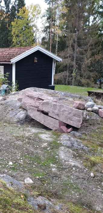 Stor röd stenbumling med lagerstruktur på en tomt med en svart stuga i bakgrunden.