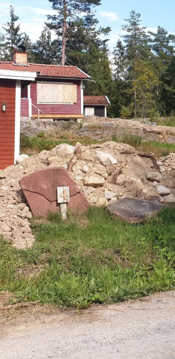 Stor röd stenbumling med skivliknande lager framför en tomt med hus och träd i bakgrunden.