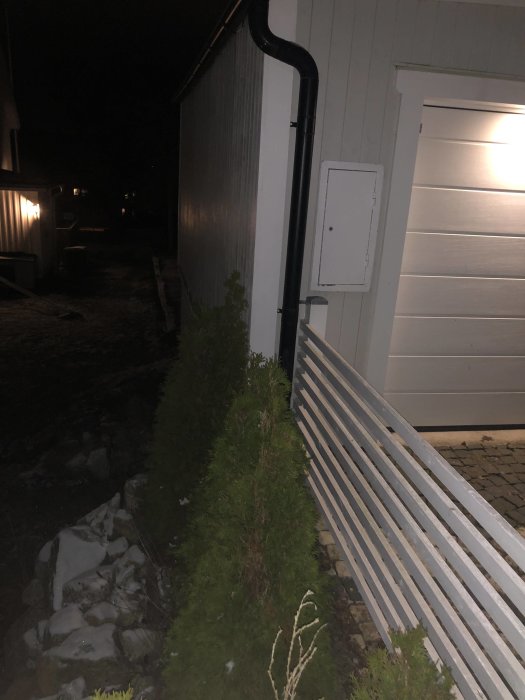 Garagegavel vid tomtgräns med ett staket och thujor planterade intill husväggen på kvällen.