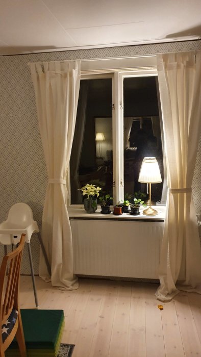 Ett fönster med vita draperade gardiner, enkelt fönsterfoder, en bordlampa och krukväxter på fönsterbrädan, med parkettgolv.