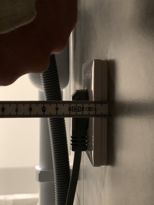 Måttband som mäter avståndet mellan en vägg och en elektrisk sladd med stickpropp framför ett uttag.