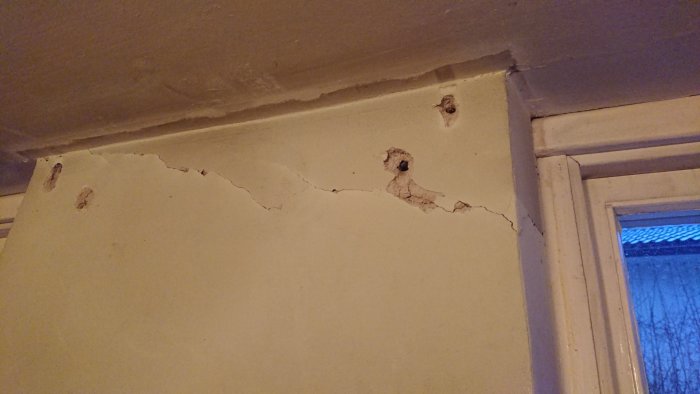 Sprickor längs övre delen av en källarvägg där träpanel tagits bort, nära taket och ett fönster.