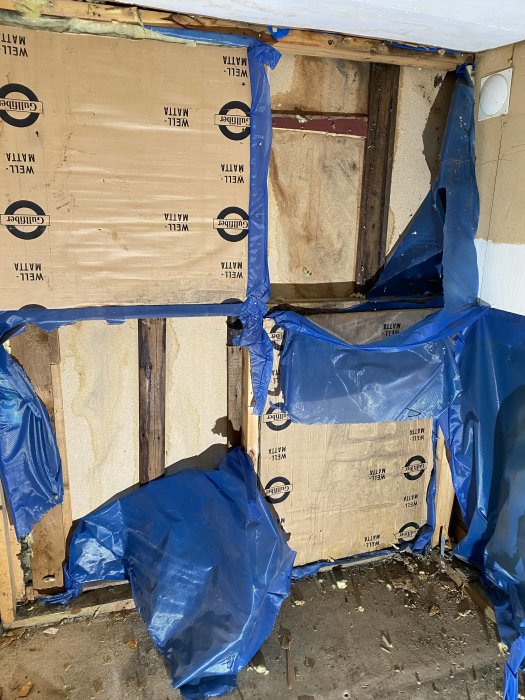 En delvis riven vägg med blå presenning och exponerade spånskivor och reglar.