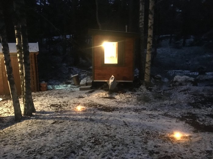 Bastu i skogsmiljö upplyst av inomhusbelysning och utomhuslampor med snötäckt mark.