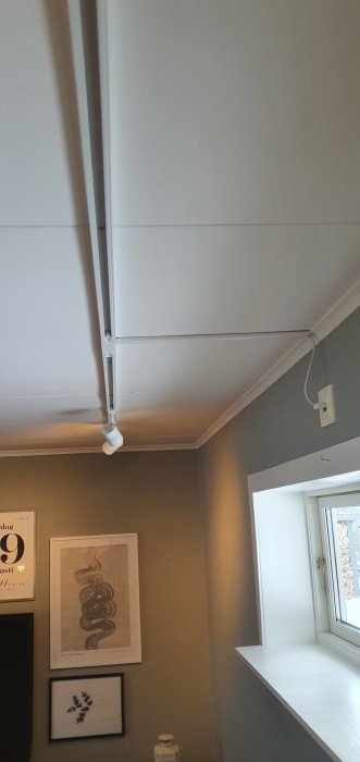 Interiör bild av ett rum med grå väggar, vitmålade dörr- och fönsterkarmar, en lampa som hänger från en kabelkanal och konst på väggen.