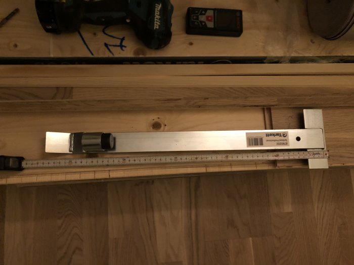 Liggande vinkelhake mot träbakgrund med tumstock och borr i bakgrunden.