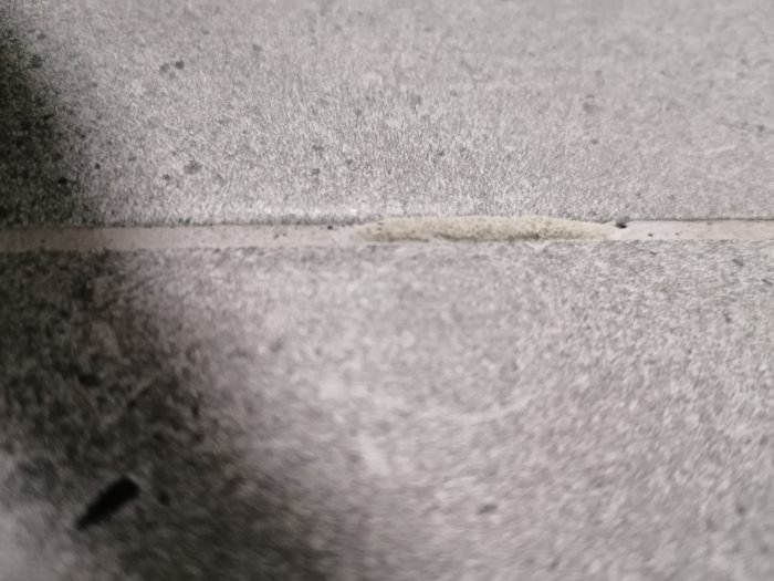 Hård, förhöjd fläck i fog mellan två gråa plattor på golv som ser ut som en ojämn ansamling material.