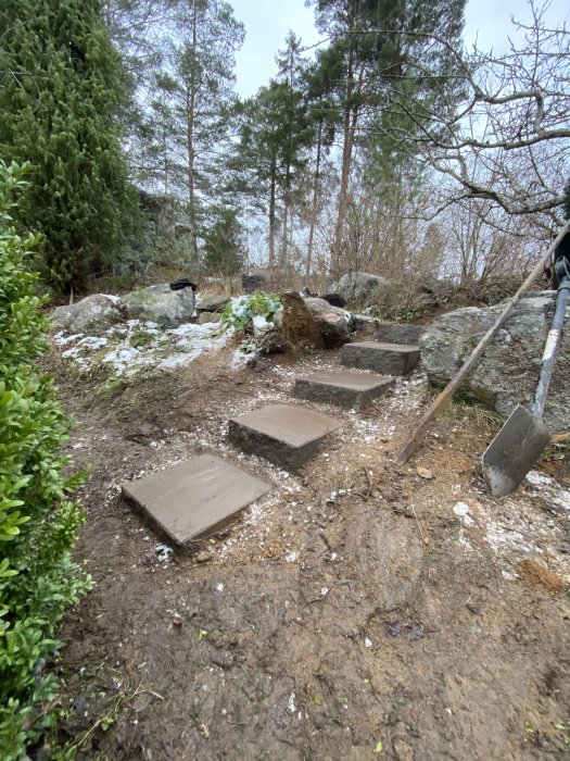 Nyanlagd trappa med fem betongsteg på en lerig backe i en skogsmiljö, med spade i förgrunden.