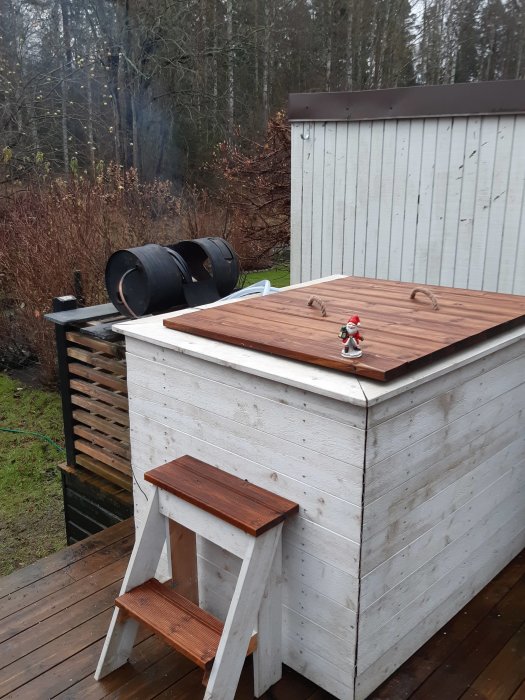 Vedeldad badtunna i trädgården med stegpall framför och julpyntad figur på lock, omgiven av natur.