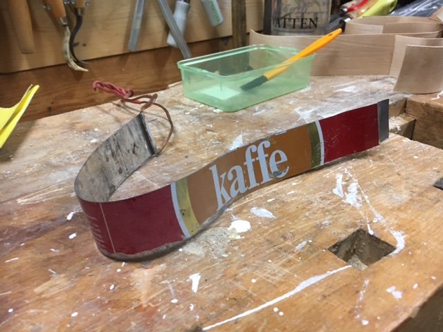 Böjd metallremsa med texten "kaffe" på ett slitet arbetsbord, använd som verktyg för att forma trädelar till ett instrument.