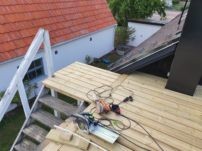 Vy över en nyligen byggd träterrass med verktyg och byggmaterial, vid sidan av en vit trätrappa och ett hus med tegeltak.