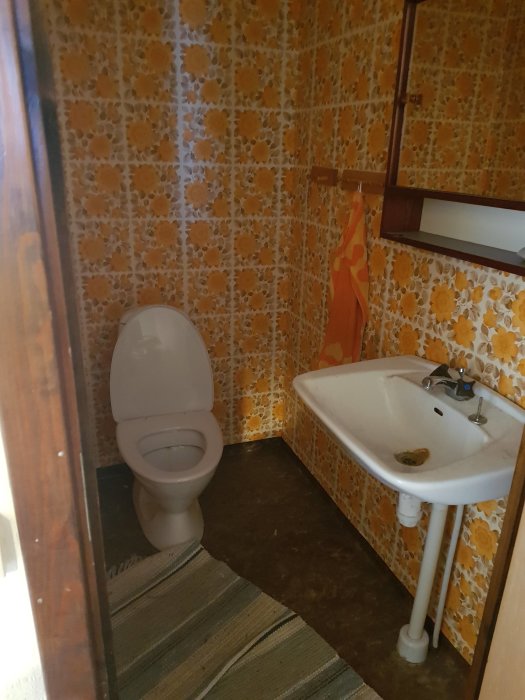 Gammalt badrum med orange blommig kakel, vit toalett och handfat redo för renovering.