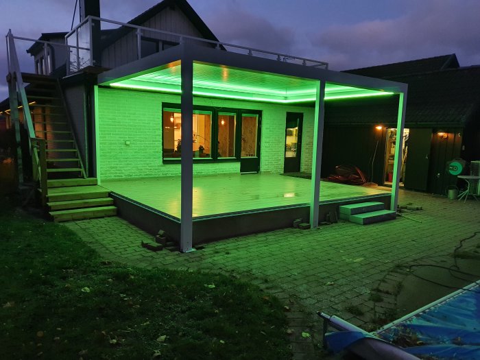 Kvällsbild av en nyligen monterad pergola med grön LED-belysning över en träterrass intill ett hus.