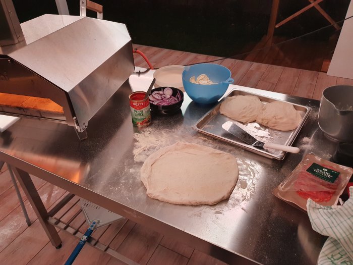 Förberedelse av pizza med deg, tomatsås och ingredienser på ett rostfritt stål köksbord med en pizzaugn.