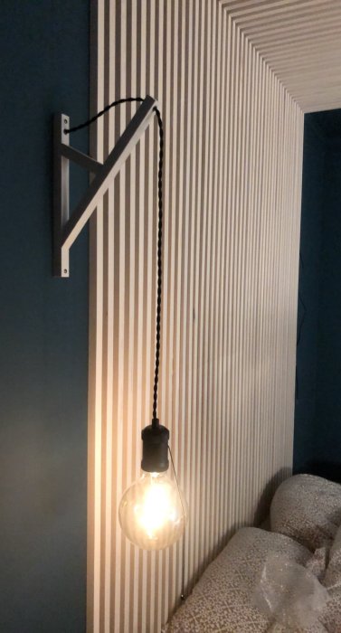 Sänggavel med vertikala strippar och svart vägglampa med glödlampa över sängen.