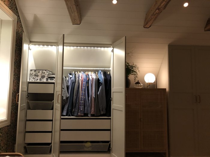 Renoverat sovrum med öppen garderob full av hängande kläder, lådhurts och en bordslampa.