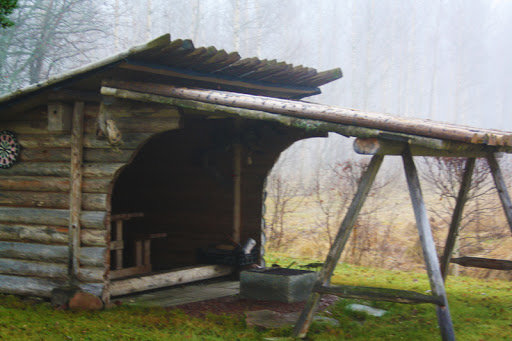 En traditionell slogbod i Dalarna med tak över en eldstad och öppen konstruktion bland träd i dimma.
