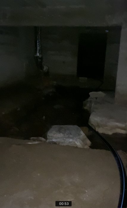 Dimly lit crawl space with standing water, rusted pipes, and concrete underside of floor joist.