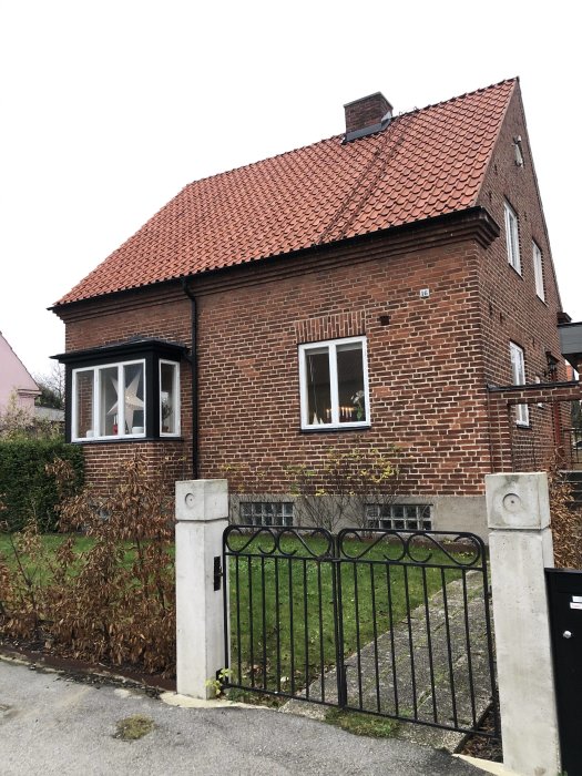 Bild på ett tegelhus från 50-talet med rött tegeltak, svart smidesstaket och vissna buskar framför.
