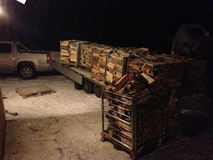 Släpvagn lastad med vedpallar på snöig mark nattetid, redo för transport med pallyftare.