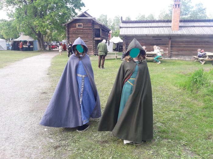 Två personer i medeltida klänningar och mantlar på en historisk marknad.