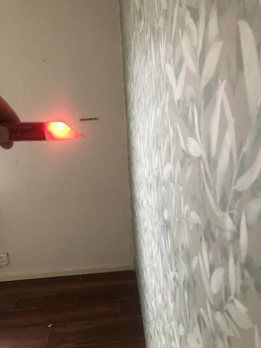 En laseravståndsmätare riktar en röd laserpunkt mot en vit vägg med tapet med bladmönster.