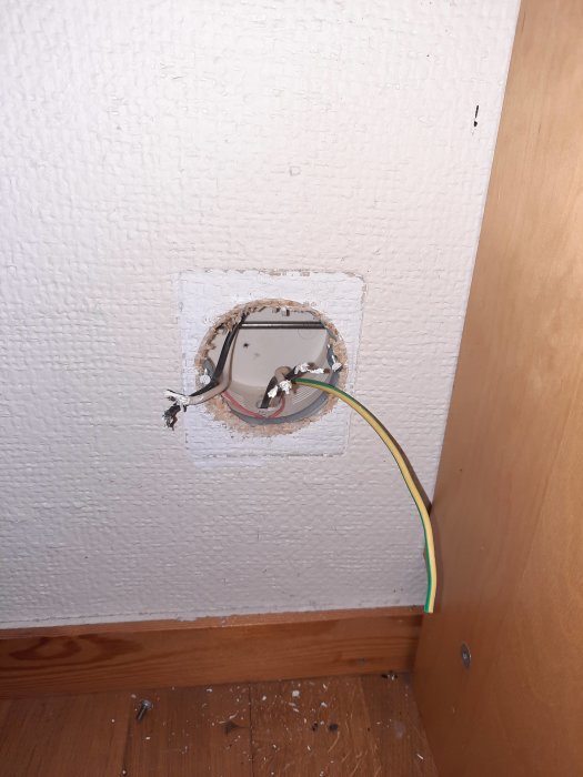 Öppet gammalt vägguttag under byte med synliga ledningar och jordkabel, på en vit vägg och parkettgolv.