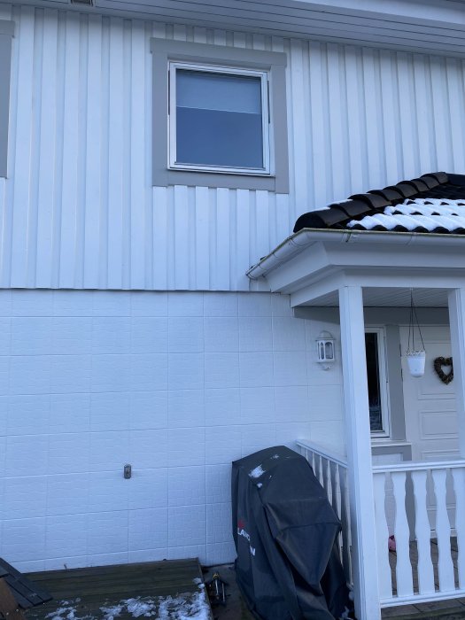 Vitt hus med liggande panel, ett fönster, snötäckt tak, och en ingångsparti med veranda samt en grill under skyddsöverdrag.