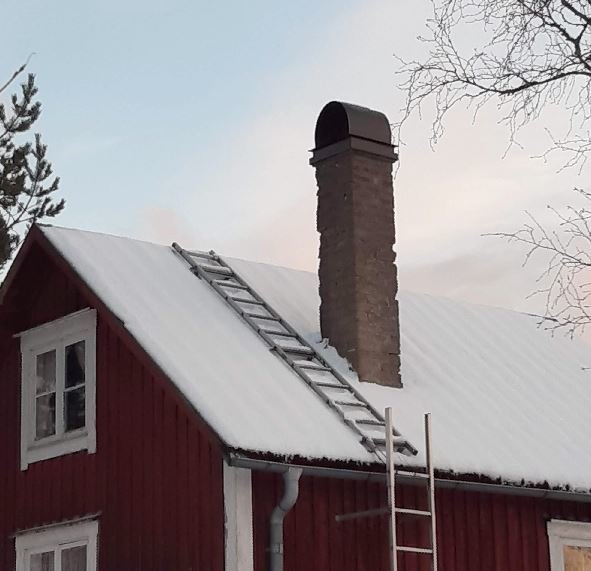 Gammal skorsten på snötäckt tak av rött hus med stege lutad mot taket.