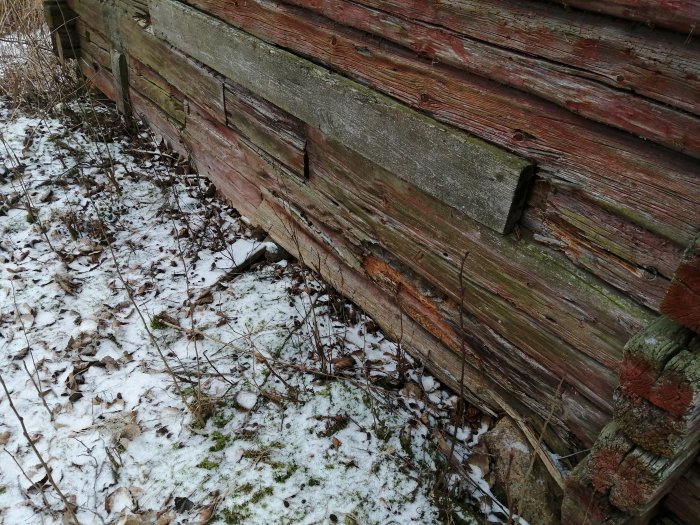 Väderbiten träfasad på en äldre byggnad med synliga tecken på förfall, omgiven av snö och löv.