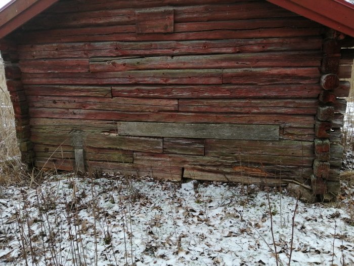 Slitet trävägg på rödfärgat timmerhus med synliga tecken på förfall och snöfläckar på marken.