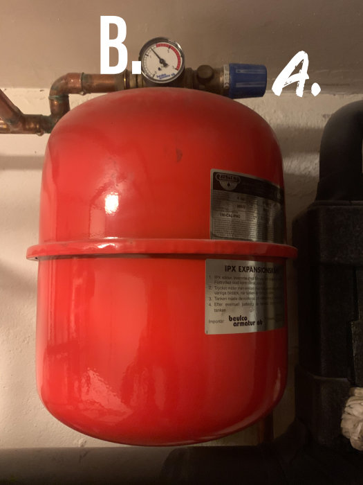 Röd expansionskärl för varmvatten med påtryckta etiketter, omgiven av rör, utrustad med manometer (B.) och ventiler (A.).