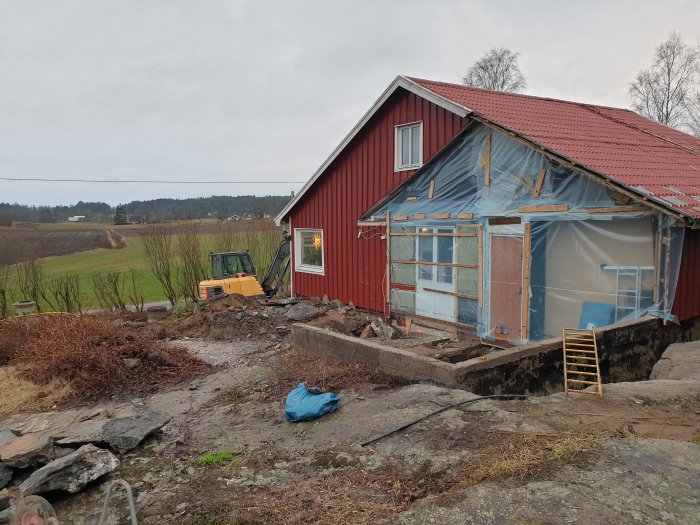 Ett rött hus under renovering med en provisorisk plasttäckt väderskydd på gaveln och en grävmaskin bredvid.